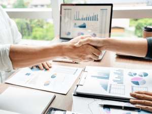 Two people shaking hands over a desk with business charts and a laptop.