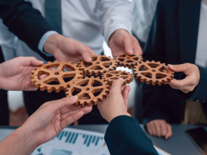 Several hands connecting wooden gears above a table with documents.